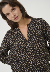 Blouse noire avec un motif léopard en beige et crème. Présente un décolleté en V et des manches longues. Tissu léger et texturé.