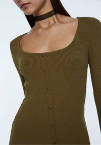 Robe côtelée vert olive avec un décolleté carré, des manches longues et une rangée de boutons sur le devant. Elle présente un tissu texturé et une silhouette ajustée.