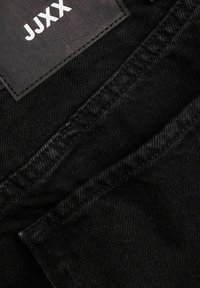 Κοντινή εικόνα μαύρου τζιν υφάσματος που δείχνει το ράψιμο της τσέπης και μια μαύρη ετικέτα με λευκό κείμενο "JUXx".