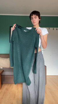 Sweater verde canelado de mangas longas com decote redondo, apresentando fendas laterais, segurado por uma pessoa vestindo uma blusa branca e calças cinzas.