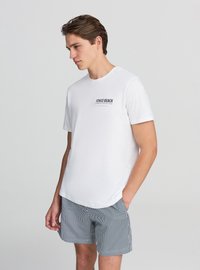 T-shirt bianca a maniche corte con grafica nera "Venice Beach", abbinata a pantaloncini a righe blu e bianche. Design semplice e vestibilità comoda.