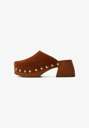 Sabots en daim marron avec un épais talon en bois. Présente des clous dorés le long du bord avant et des détails cousus sur le dessus.