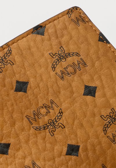 Κοντινό πλάνο υφής από ταμπά δέρμα με μαύρα λογότυπα MCM και διαμάντια, που δείχνει λεπτομερή ραφή στη γωνία.