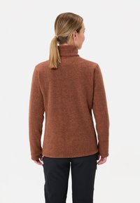 Gestructureerde bruine fleecejack met een hoge kraag en duimgaten, gecombineerd met donkere broek. De achterkant toont een eenvoudig, functioneel ontwerp.