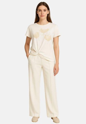 Crèmewitte T-shirt met een knoop aan de voorkant, voorzien van een subtiele bloemenprint en het woord "BLOOM." Gecombineerd met crèmekleurige broek met wijde pijpen.