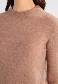 Brązowy sweter dziany z okrągłym dekoltem, miękką fakturą i dopasowanym krojem. Tkanina ma delikatny połysk i drobne sploty.
