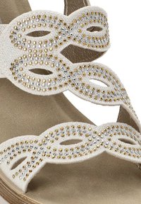 Sandalia blanca con tiras superpuestas adornadas con strass de oro y plata, sobre una plantilla acolchada color beige con un acabado texturizado.