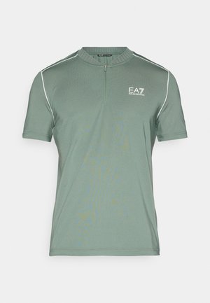 Camiseta de manga corta con cuello con cremallera en un color verde apagado. Presenta costuras contrastantes claras y un logo en relieve en el pecho izquierdo.