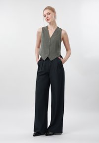 Jeune femme debout les mains dans les poches, portant un gilet gris sans manches boutonné, un pantalon large noir et des chaussures noires pointues sur fond blanc.