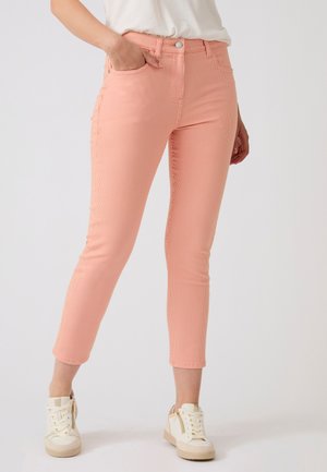 Pantaloni aderenti color pesca a costine indossati con sneakers bianche e camicia bianca, mostrati su una persona in piedi su uno sfondo semplice.