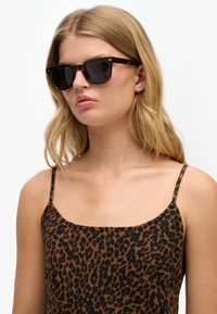 Gafas de sol con montura de carey y lentes oscuros. Llevadas con una camiseta sin mangas de estampado de leopardo marrón con tirantes finos.