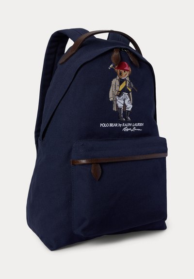 Rucsac albastru marin cu mâner din piele maro și detalii de fermoar, având brodat pe față un urs în costum de călărie care ține un ciocan de polo.