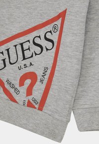 Sweatshirt gris avec un graphique triangulaire rouge portant "GUESS® U.S.A." et des détails en texte, incluant "WASHED," en noir. Texture de tissu lisse.