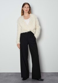Cardigan aperto fluffy di colore bianco sporco, realizzato in tessuto testurizzato, abbinato a pantaloni larghi neri a vita alta e scarpe nere con punta.