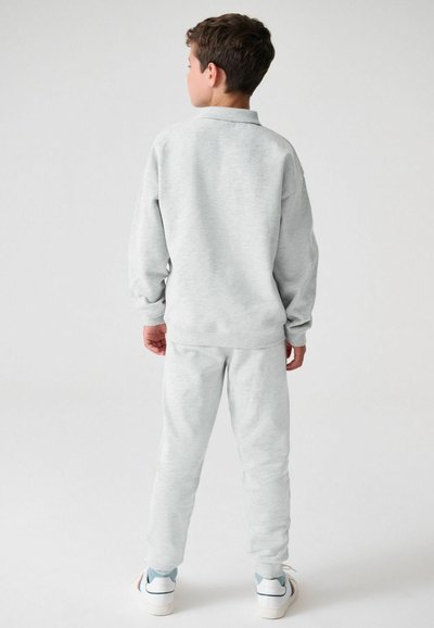 Šviesiai pilka flisinė sportinė apranga, susidedanti iš džemperio ir kelnių, pasižyminti laisvu siluetu, virbalų apykakle ir juosmeniu, minkštu tekstūru ir paprastu dizainu.