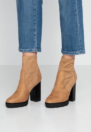 Tan semskede ankelstøvler med sorte chunky høje hæle båret med blå cropped jeans på en hvid gulv og ensfarvet gråt baggrund.