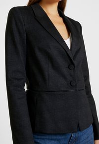 Tailored black blazer in patterned fabric with two front buttons. Features a structured collar and fitted waist, complemented by long sleeves.
