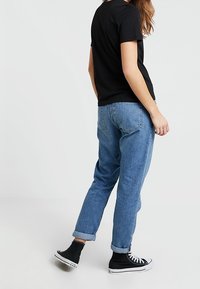 Person iført opkrøllede blå jeans, sorte sneakers med højt skaft og en sort kortærmet t-shirt, der står mod en ensfarvet lys baggrund.