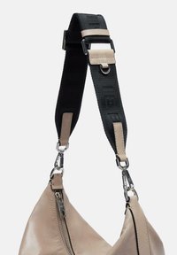 Beżowa skórzana torba o teksturowanym wykończeniu, z czarnym paskiem z logo, srebrnymi elementami i zamkiem błyskawicznym.