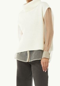 Vrouw in een witte mouwloze gebreide turtleneck over een doorschijnende beige blouse met lange mouwen en donkergrijze jeans, met haar hand aan de zijkant.