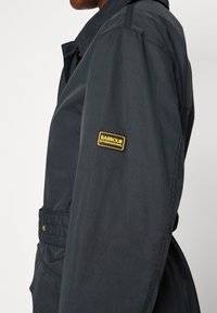 Vue latérale d'une personne portant une veste sombre avec un patch jaune et noir "Barbour International" sur le haut de la manche.