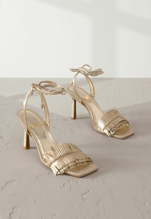 Sandale cu toc înalt aurii, cu vârf pătrat, având un design cu franjuri pliați pe curea și curele legate la gleznă. Textură metalică uniformă.