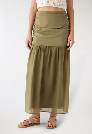 Vista dalla vita ai piedi di una persona che indossa una gonna lunga a strati verde oliva e sandali piatti beige con cinturini alla caviglia, in piedi contro uno sfondo bianco.