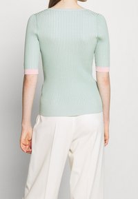 Haut en maille côtelée vert clair avec un col rond dégagé et des manches courtes ornées de poignets roses. Le design est ajusté et lisse, mettant en valeur la texture.