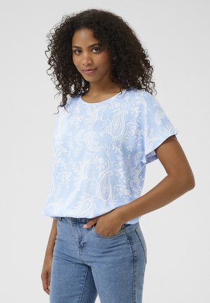 Light blue short-sleeve blouse with a white paisley pattern, round neckline, and a loose fit, paired with denim jeans. Smooth texture.