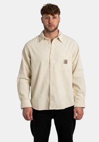 Camisa de pana beige con cierre de botones al frente, mangas largas y un bolsillo en el pecho con un parche de logo. Patrón acanalado texturizado, ajuste relajado.