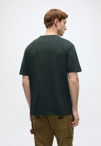  Σκούρο πράσινο βαμβακερό κοντομάνικο μπλουζάκι με στρογγυλή λαιμόκοψη, επίπεδες ραφές και χαλαρή εφαρμογή. Φορεμένο με ανοιχτό καφέ παντελόνι καμφοράς.