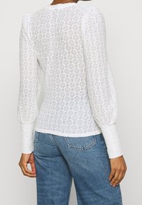 Bílý dlouhý rukáv top vyrobený z texturované krajky s nafouknutými rameny a kulatým výstřihem, v kombinaci s modrými džínami.