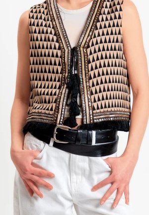 Gilet senza maniche con motivo a triangoli neri, rifiniture metalliche e nappine, indossato sopra un top chiaro, abbinato a pantaloni bianchi e una cintura nera.