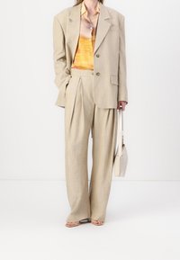 Veste de costume beige surdimensionnée avec un bouton, accompagnée de pantalons plissés assortis. Chemise à motifs orange vif et jaune. Sac à main léger.