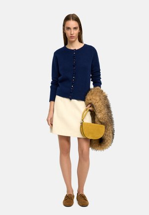 Kobieta ubrana w granatowy sweter na guziki i białą spódnicę, trzymająca musztardowo-żółtą torebkę oraz brązowy futrzany płaszcz.