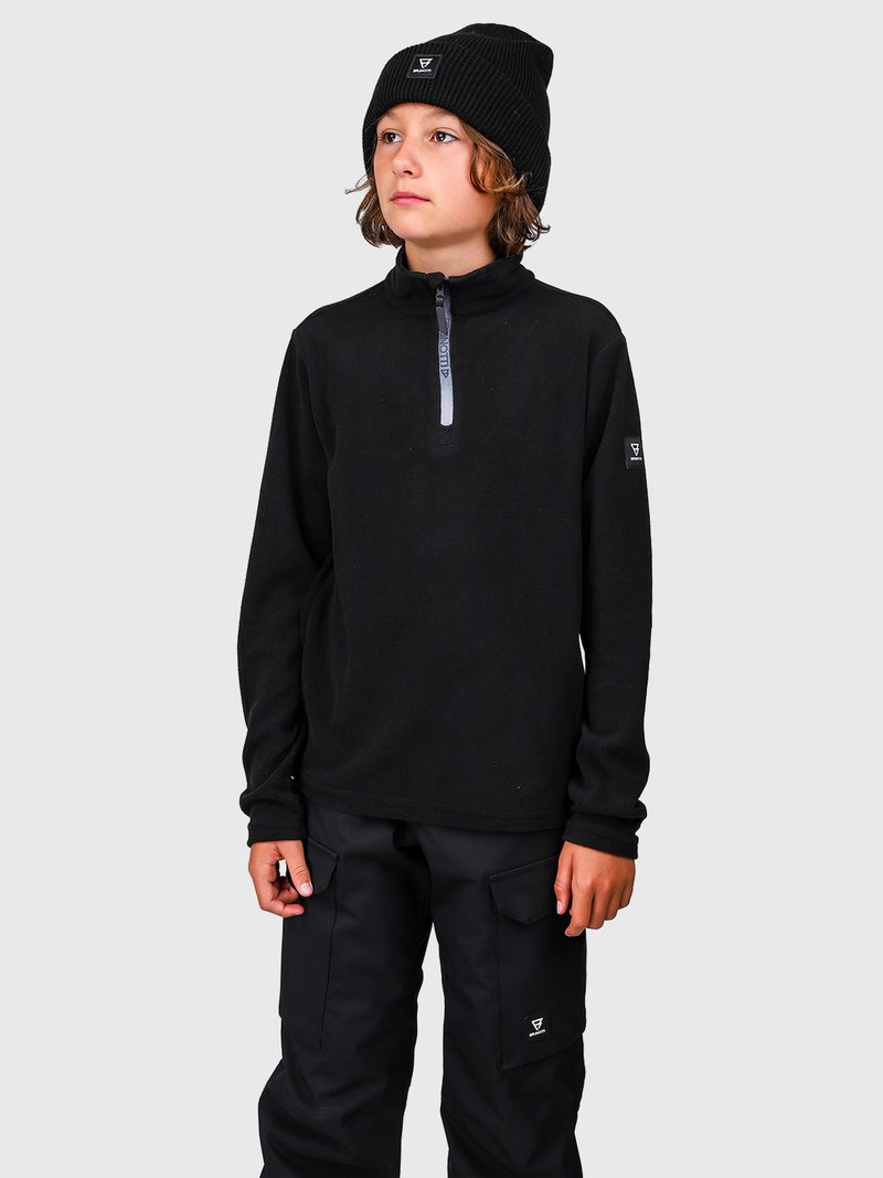 Bambino con un berretto nero, un fleece nero con zip e pantaloni cargo neri, in piedi contro uno sfondo grigio semplice, che guarda di lato.