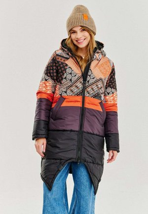 Padded long coat with a vibrant patterned design in black, orange, and brown. Features a hood and a split hem, worn over wide-leg jeans.