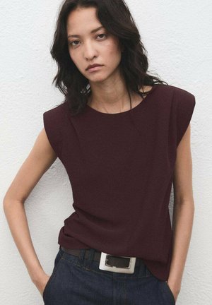 Young woman with dark wavy hair wears sleeveless dark burgundy top and blue jeans with white belt, standing against textured white wall.