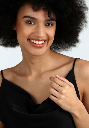 Black satin top with a cowl neckline, showcasing a hand wearing a gold band ring set with small clear stones. Model has curly hair.