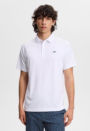 Joven con cabello rizado que lleva una camiseta polo blanca de manga corta y pantalones cortos azules con estampado, de pie frente a un fondo liso.
