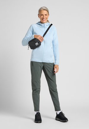 Smiling woman in light blue jacket, gray pants, and black hiking shoes stands holding small black crossbody bag against plain gray background.
