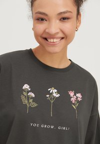 Giovane donna sorridente che indossa una maglietta grigio scuro con tre fiori ricamati e la scritta "YOU GROW, GIRL!" su uno sfondo semplice.