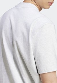 Osoba v jednoduchém světle šedém tričku s krátkým rukávem, zobrazená ze zadu a ze strany s viditelným krkem a částečně viditelnou tváří.