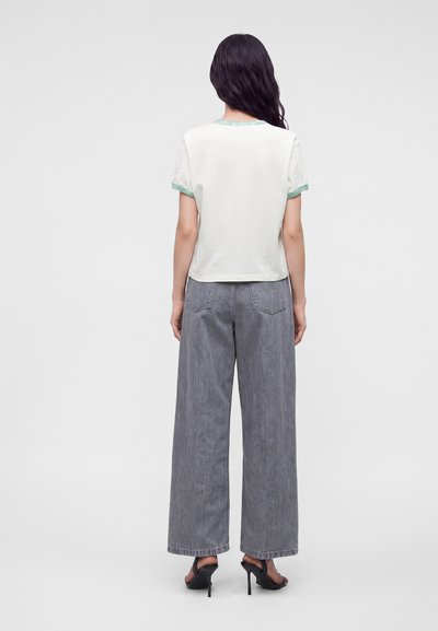 Femme aux longs cheveux foncés portant un chemisier blanc à manches courtes avec un bord vert, un jean gris à jambes larges et des sandales noires à talons hauts, debout de dos.