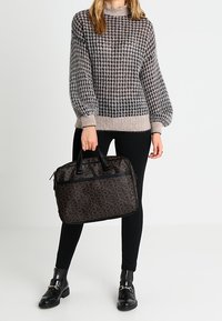 Sacoche d'ordinateur portable noire à motifs, dotée de doubles poignées, d'une texture lisse et d'une poche avant zippée. Associée à un pull gris à carreaux et à des leggings noirs.