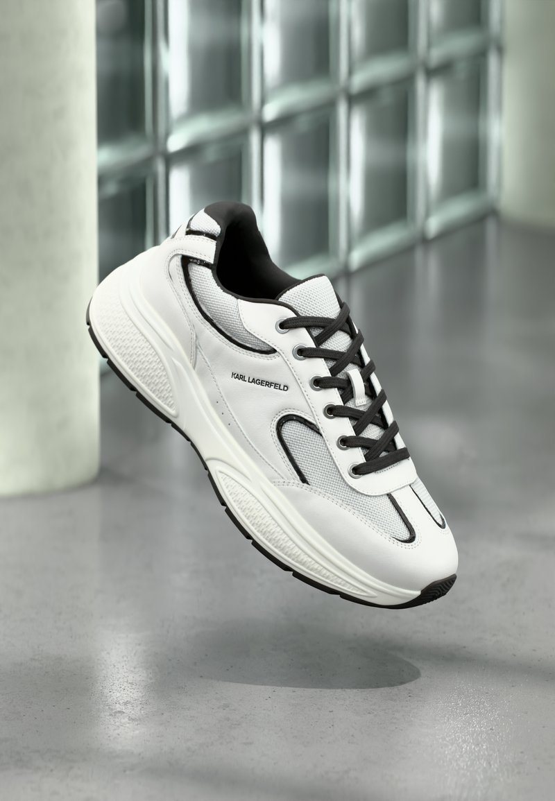 Λευκό αθλητικό παπούτσι με ύφασμα διχτυού και δερματινή επιφάνεια, μαύρες λεπτομέρειες και υφή στη σόλα. Διαθέτει κορδόνια και σήμανση στο πλάι.