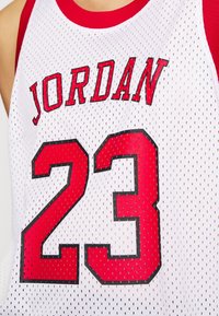 Vit baskettröja med röd kant, med "JORDAN" i röda bokstäver ovanför nummer "23" i röd och svart. Tillverkad av nätmaterial.