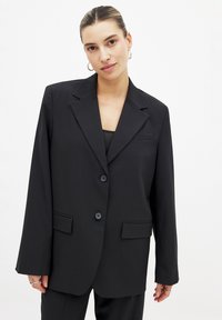 Blazer noir au design structuré, avec deux boutons, des revers à cran, des poches avant et une texture lisse. Les manches longues complètent le look.