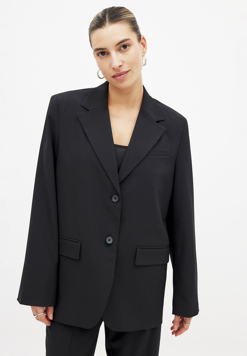 Blazer noir au design structuré, avec deux boutons, des revers à cran, des poches avant et une texture lisse. Les manches longues complètent le look.