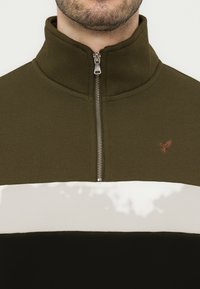 Άνδρας που φοράει πουλόβερ με φερμουάρ έως το ύψος του στήθους σε ελιά πράσινο, λευκό και μαύρο, με μικρό κεντημένο λογότυπο αετού στο στήθος.